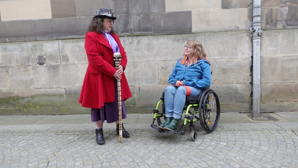 Two ladies talking outside. One is in a wheelchair, one is wearing a top hat and holding a staff.